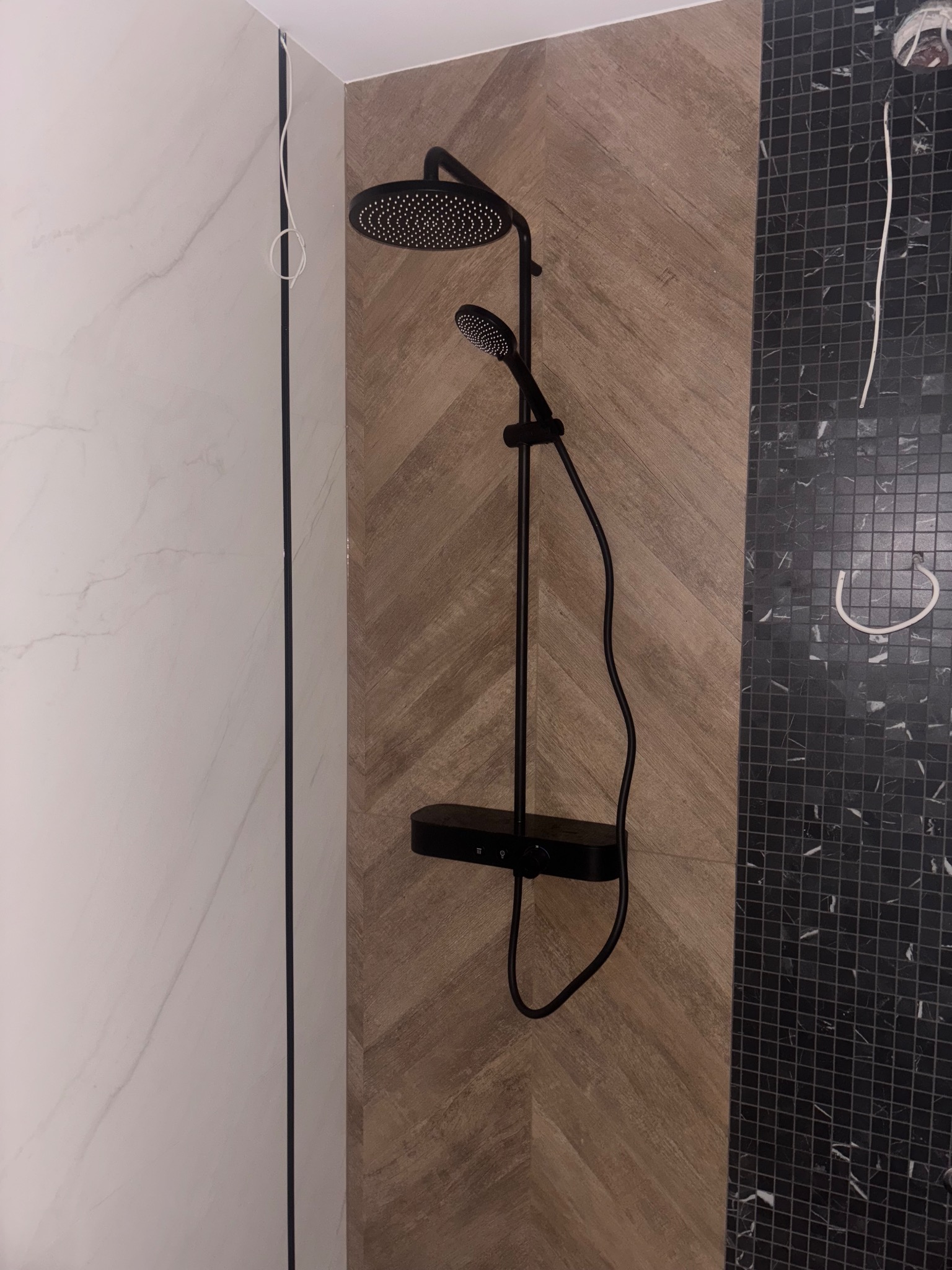 Czarny panel prysznicowy z deszczownicą i słuchawką, zamontowany na ścianie z płytek imitujących drewno, obok ściany z czarnych płytek mozaikowych.