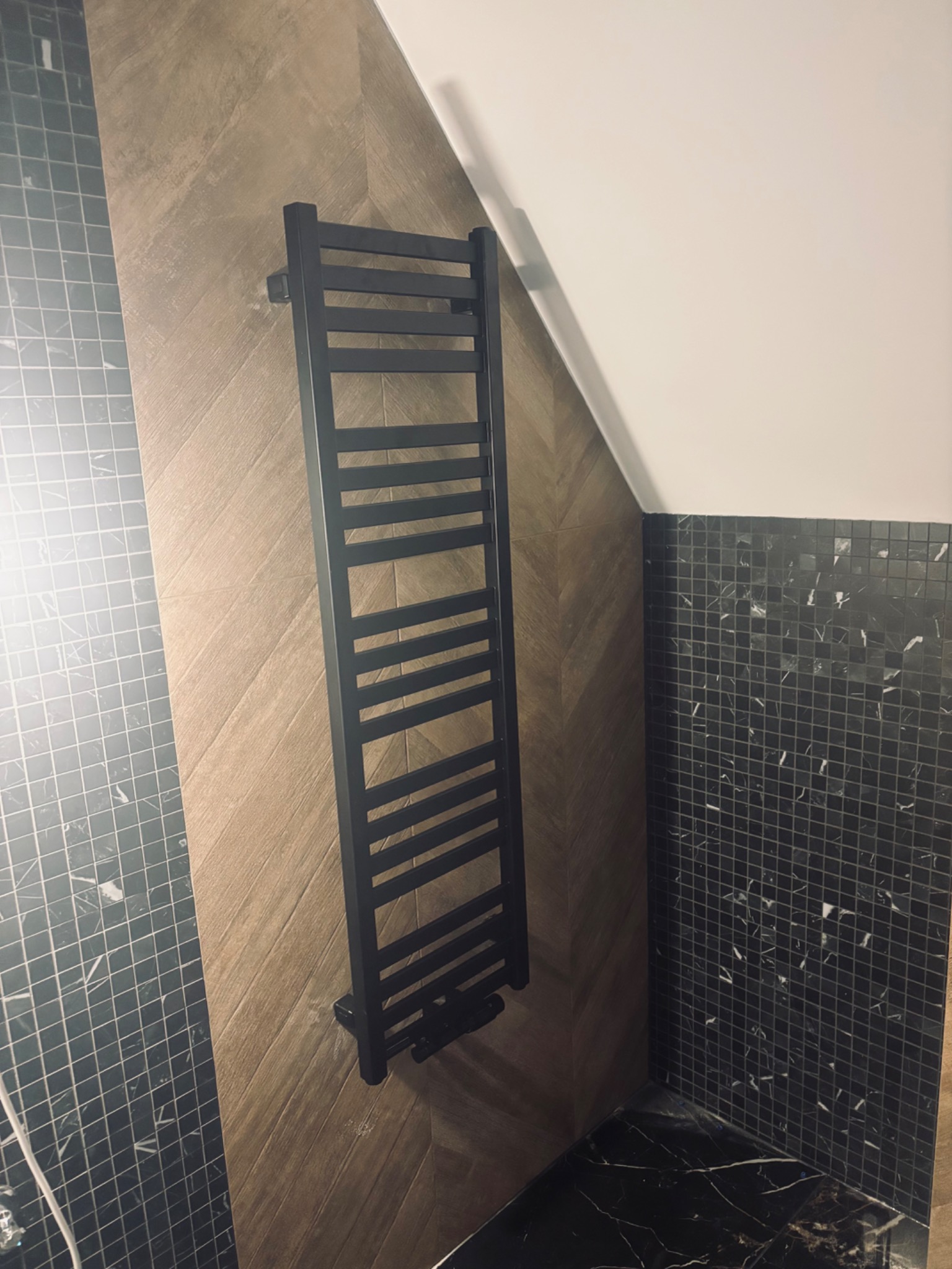 Czarny, drabinkowy grzejnik łazienkowy na ścianie z wzorem jodełki, obok ściany wyłożonej mozaiką w odcieniach czerni i szarości. Nowoczesna aranżacja łazienki.