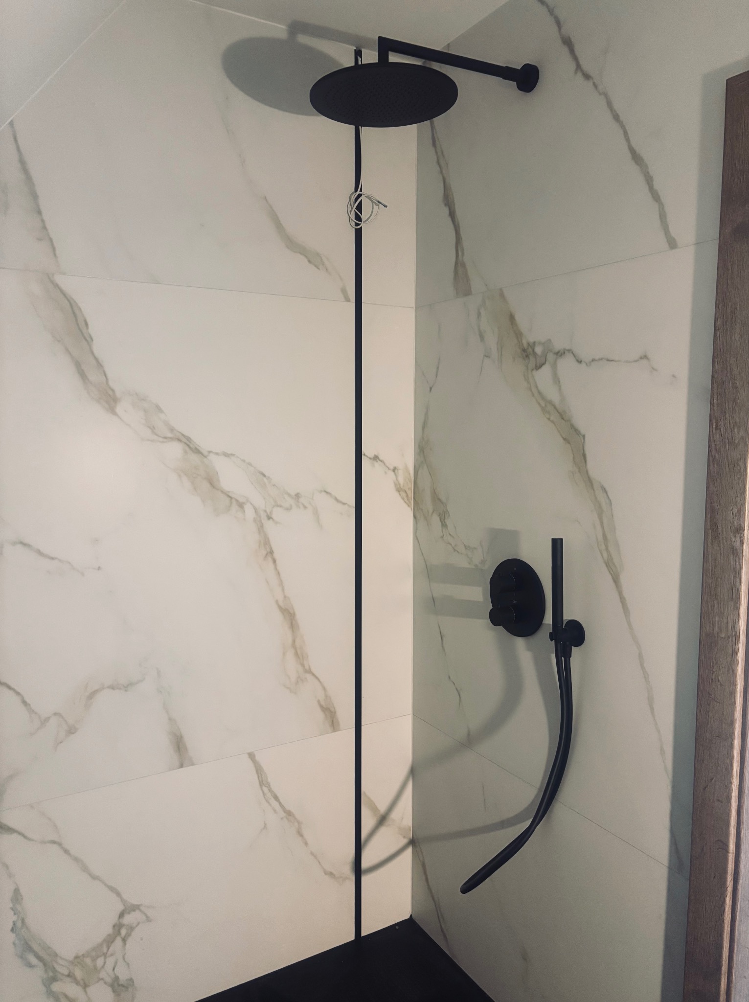 Nowoczesna kabina prysznicowa z deszczownicą w kolorze czarnym, wykończona płytkami imitującymi marmur. Minimalistyczny design, widoczne elementy instalacji.