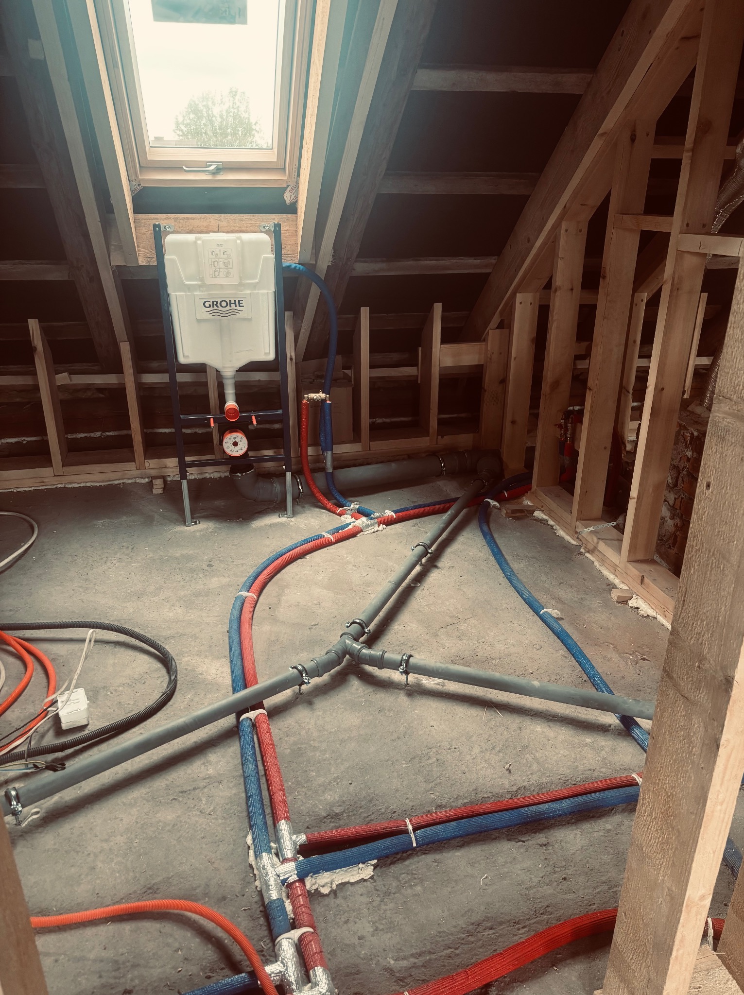 Instalacja wodna w poddaszu z oknem dachowym: rury w izolacji czerwonej i niebieskiej, szara kanalizacja, stelaż podtynkowy Grohe. Widoczne elementy konstrukcji drewnianej.