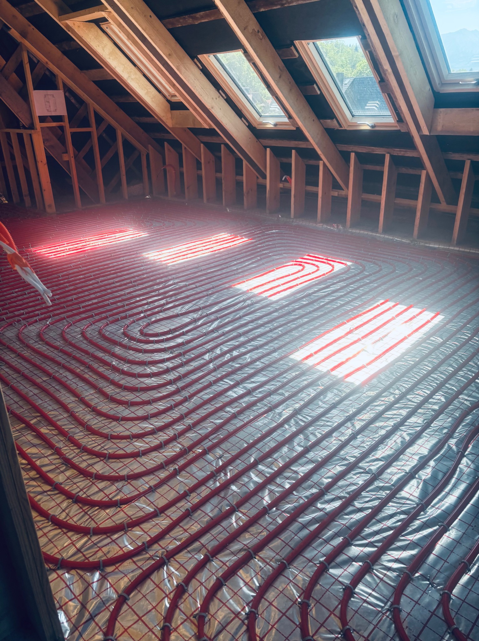 Czerwone rury ogrzewania podłogowego ułożone na folii w nowym budynku z drewnianą konstrukcją dachu i oknami dachowymi wpuszczającymi jasne światło.