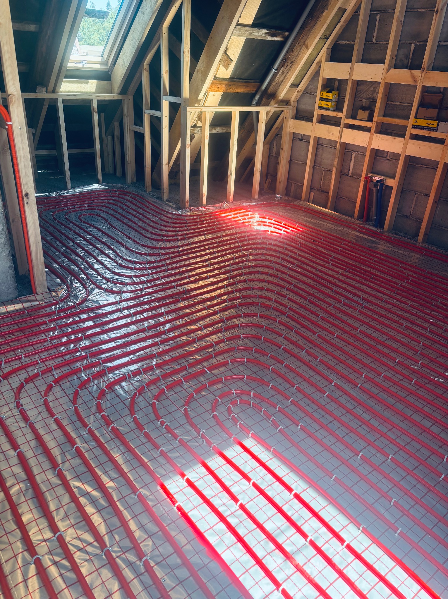 Czerwone rury ogrzewania podłogowego ułożone na folii w surowym wnętrzu poddasza z drewnianą konstrukcją i oknem dachowym. Etap instalacji przed wylewką.