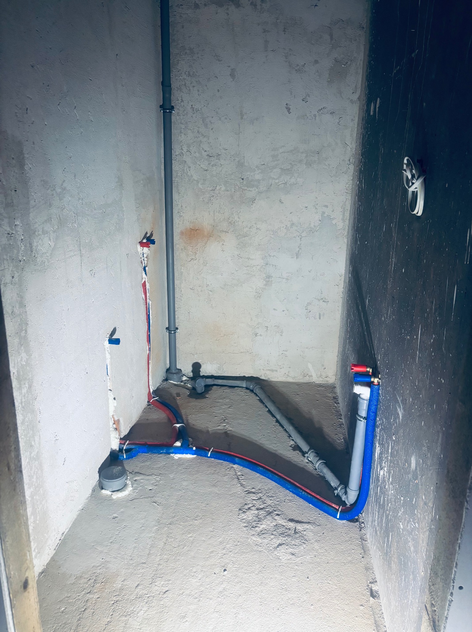 Surowe wnętrze z instalacją hydrauliczną: pionowe rury szare, poziome niebiesko-czerwone, połączone z zaworami. Ściany w stanie surowym, betonowe, w tle zaciemniona ściana.