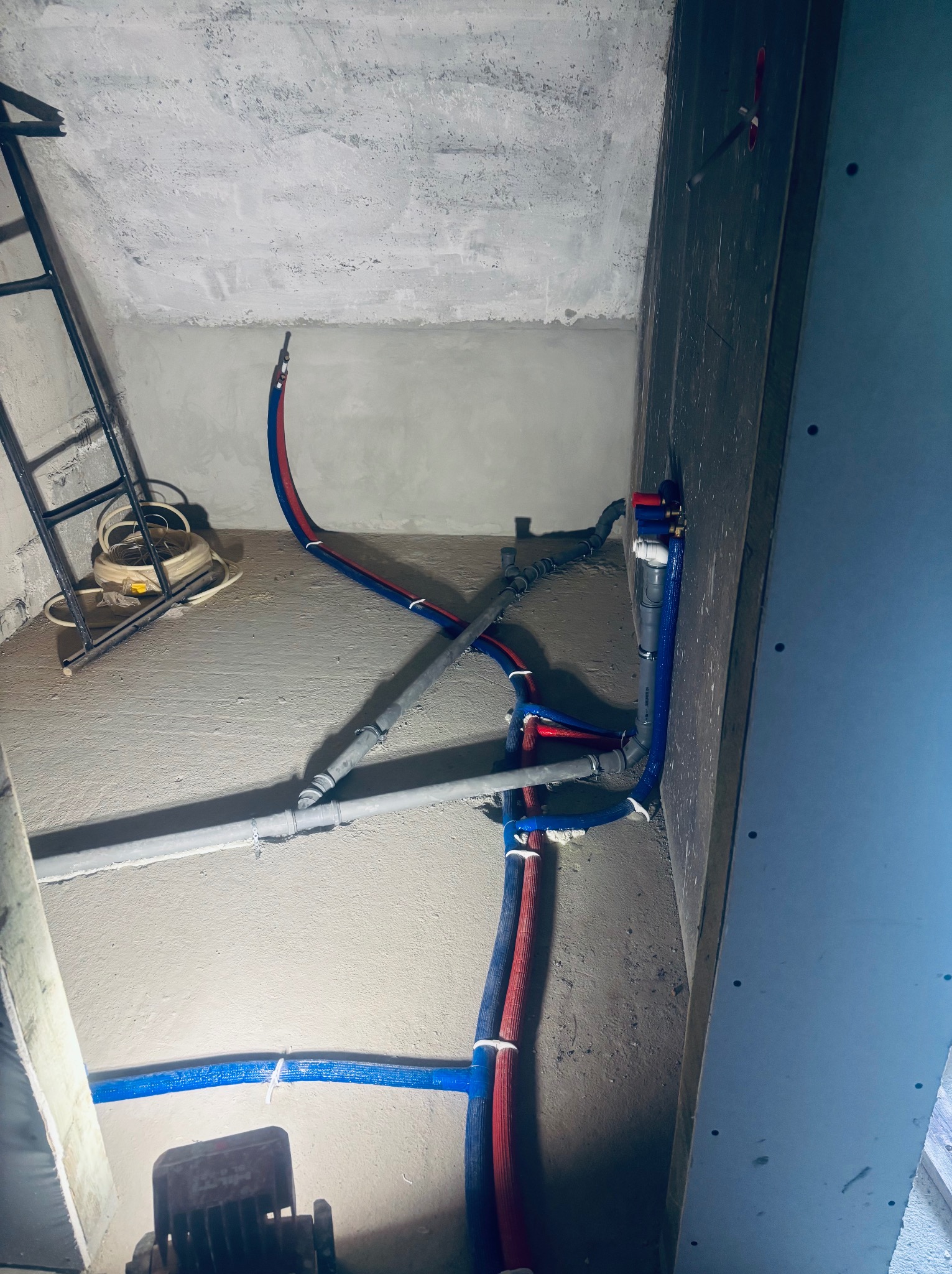 Szare i kolorowe rury instalacji wodnej ułożone w surowym wnętrzu. Widoczna drabina i przewody elektryczne. Etap prac instalacyjnych w pomieszczeniu.