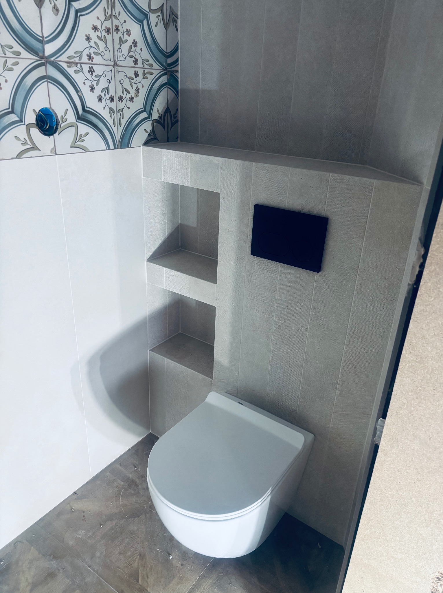 Nowoczesna toaleta podwieszana z zabudową wnękową wykończona szarą płytką imitującą kamień, akcentowana dekoracyjną płytką w stylu retro nad stelażem.