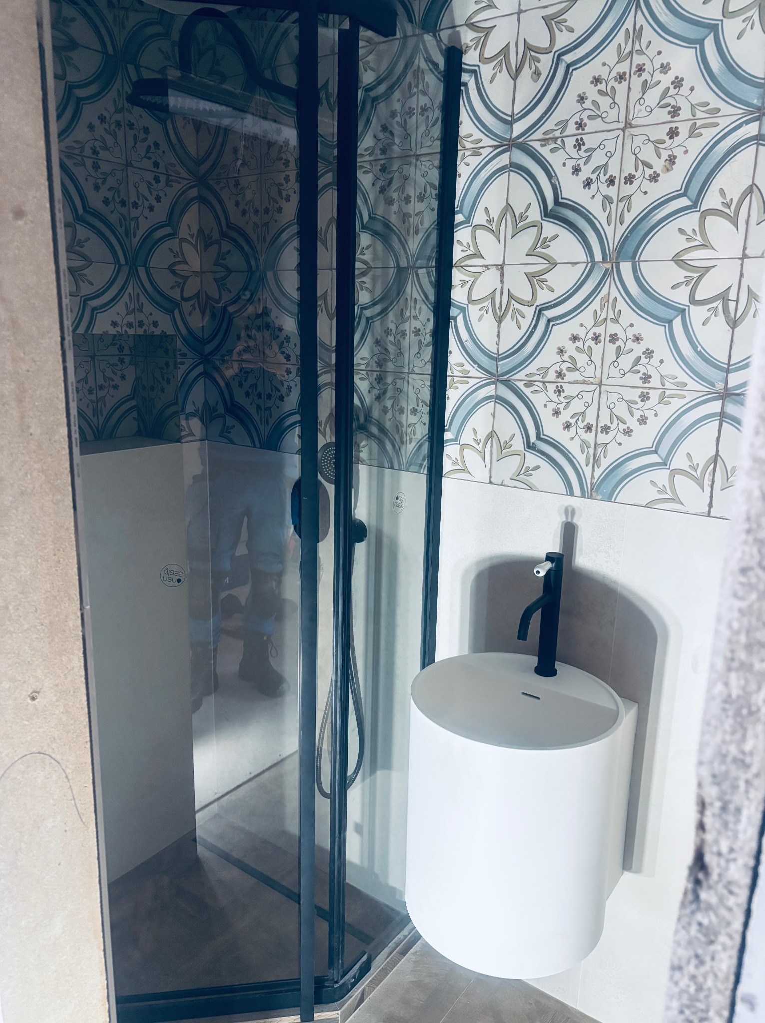 Nowoczesna łazienka z kabiną prysznicową w czarnej ramie i białą, okrągłą umywalką nablatową z czarną armaturą na tle dekoracyjnych płytek.