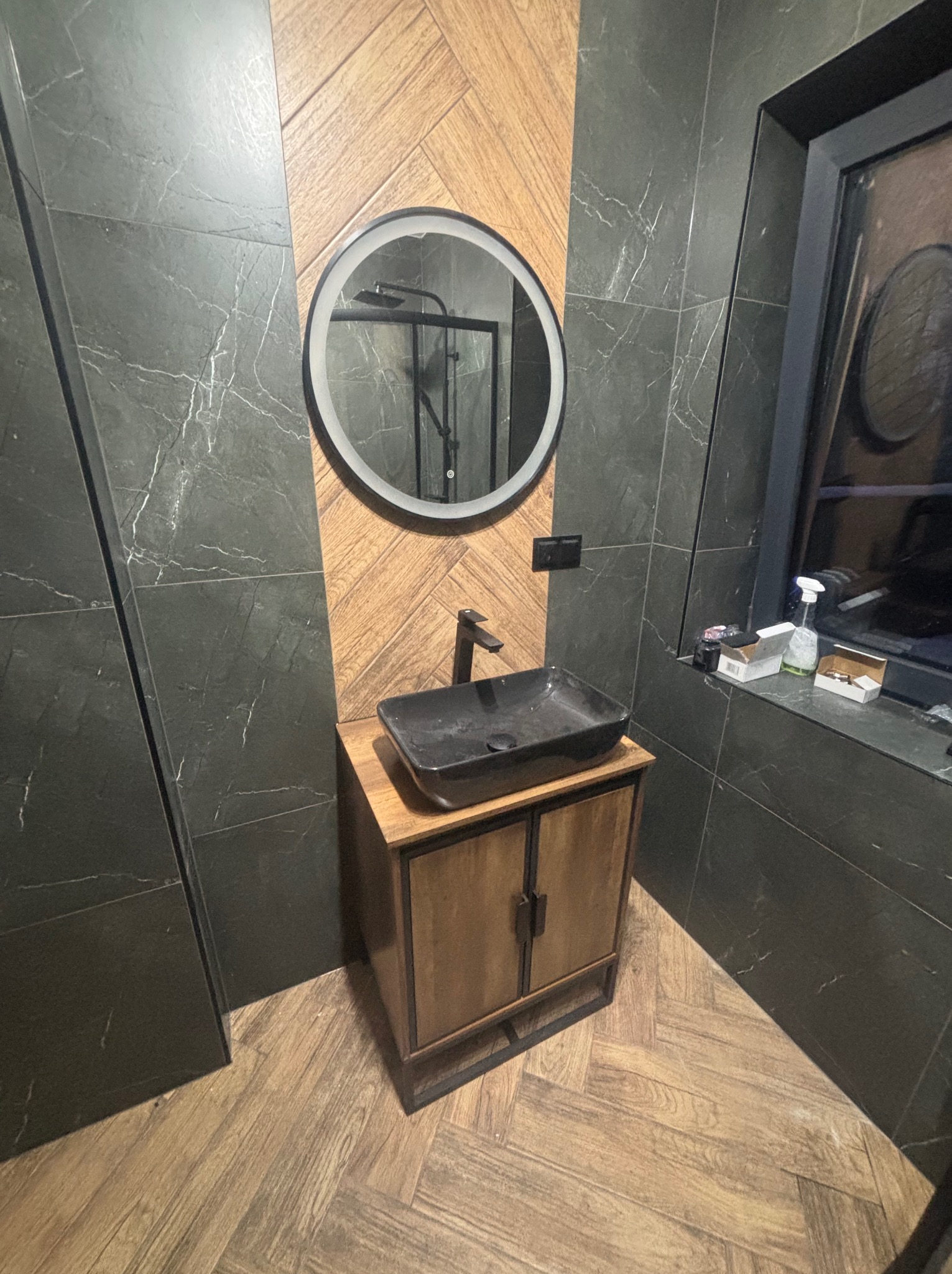 Nowoczesna łazienka z umywalką nablatową, okrągłym lustrem z podświetleniem LED i czarnymi płytkami imitującymi marmur. Drewniana podłoga w jodełkę.