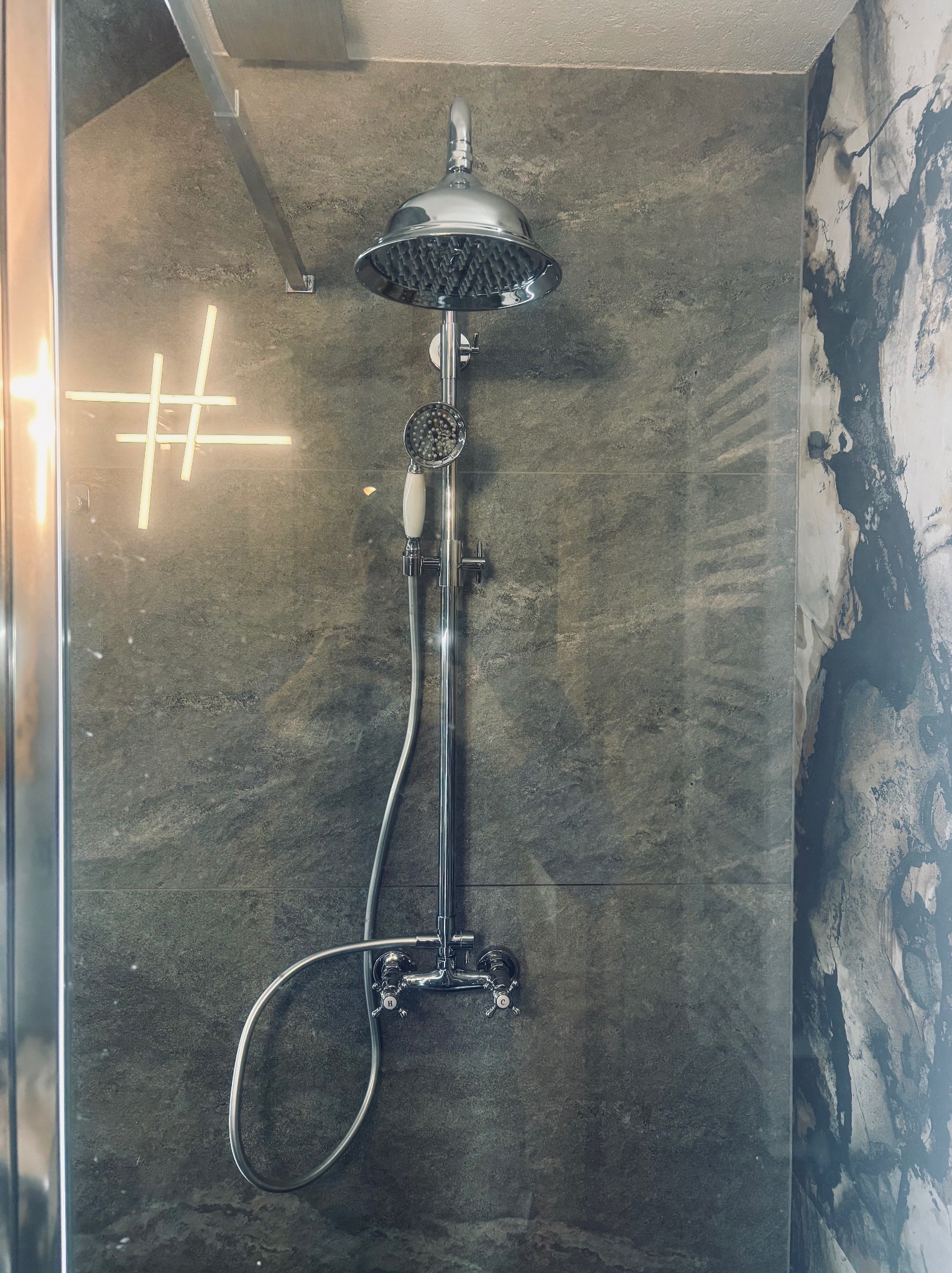 Chromowana bateria natryskowa z deszczownicą i słuchawką, zamontowana na szarej, marmurkowej ścianie w kabinie prysznicowej z dekoracyjnym oświetleniem.