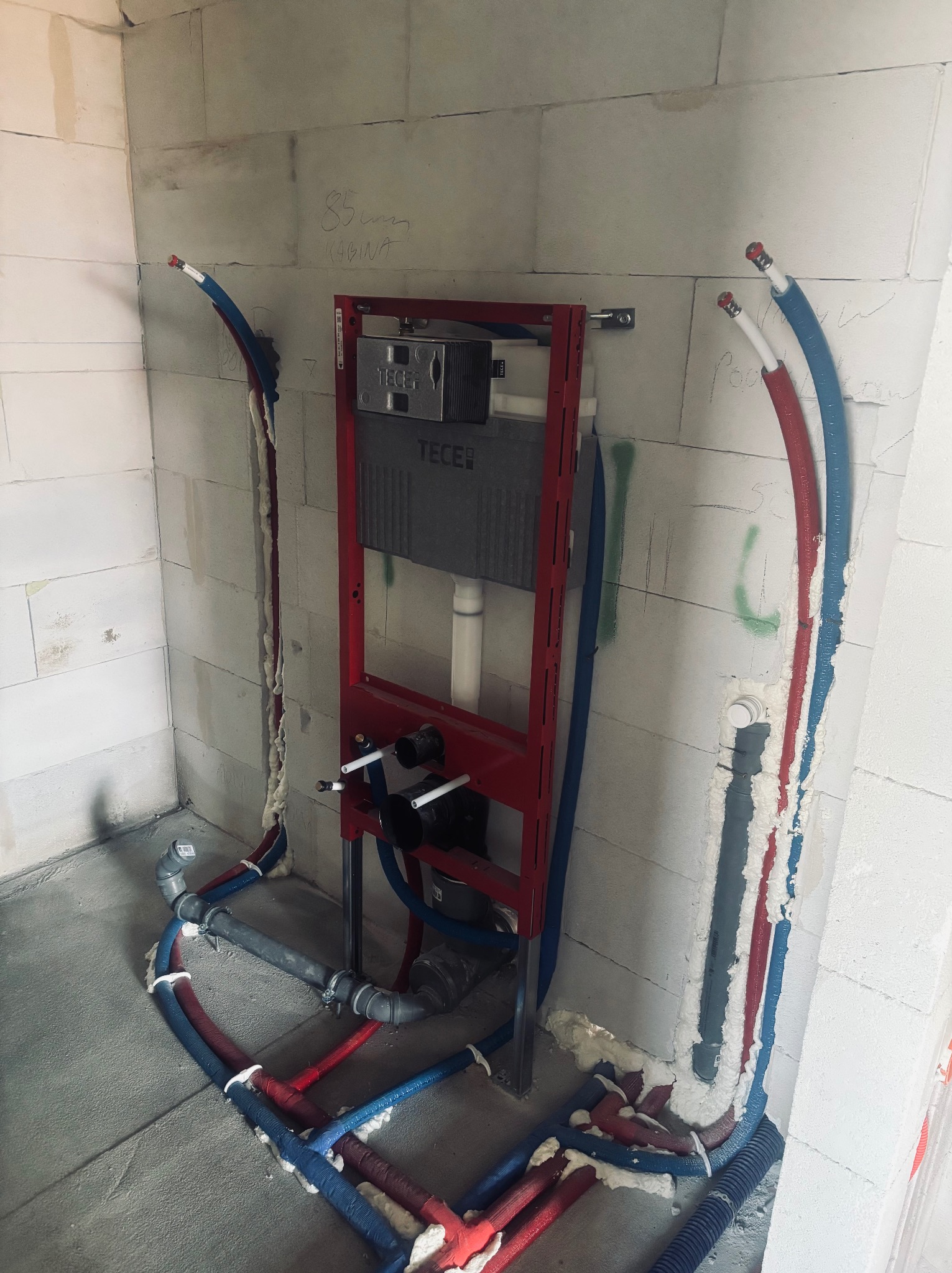 Stelaż WC TECE w czerwonej ramie, z podłączonymi rurami wodnymi w izolacji czerwono-niebieskiej, osadzony w surowej ścianie z betonu komórkowego.