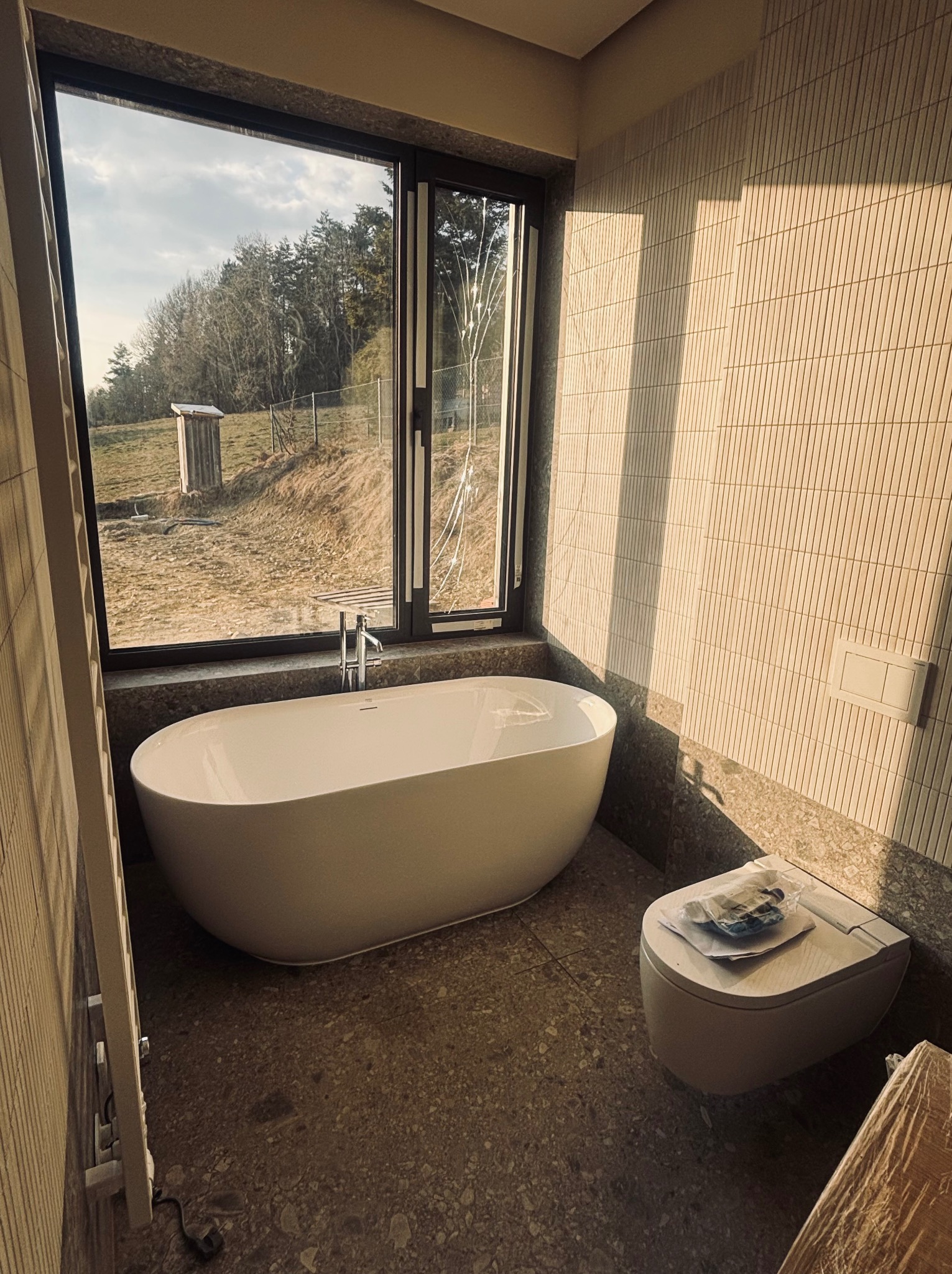 Nowoczesna łazienka z wolnostojącą wanną przy oknie z widokiem na krajobraz. Umywalka i toaleta w minimalistycznym stylu. Ściany wyłożone beżową mozaiką.