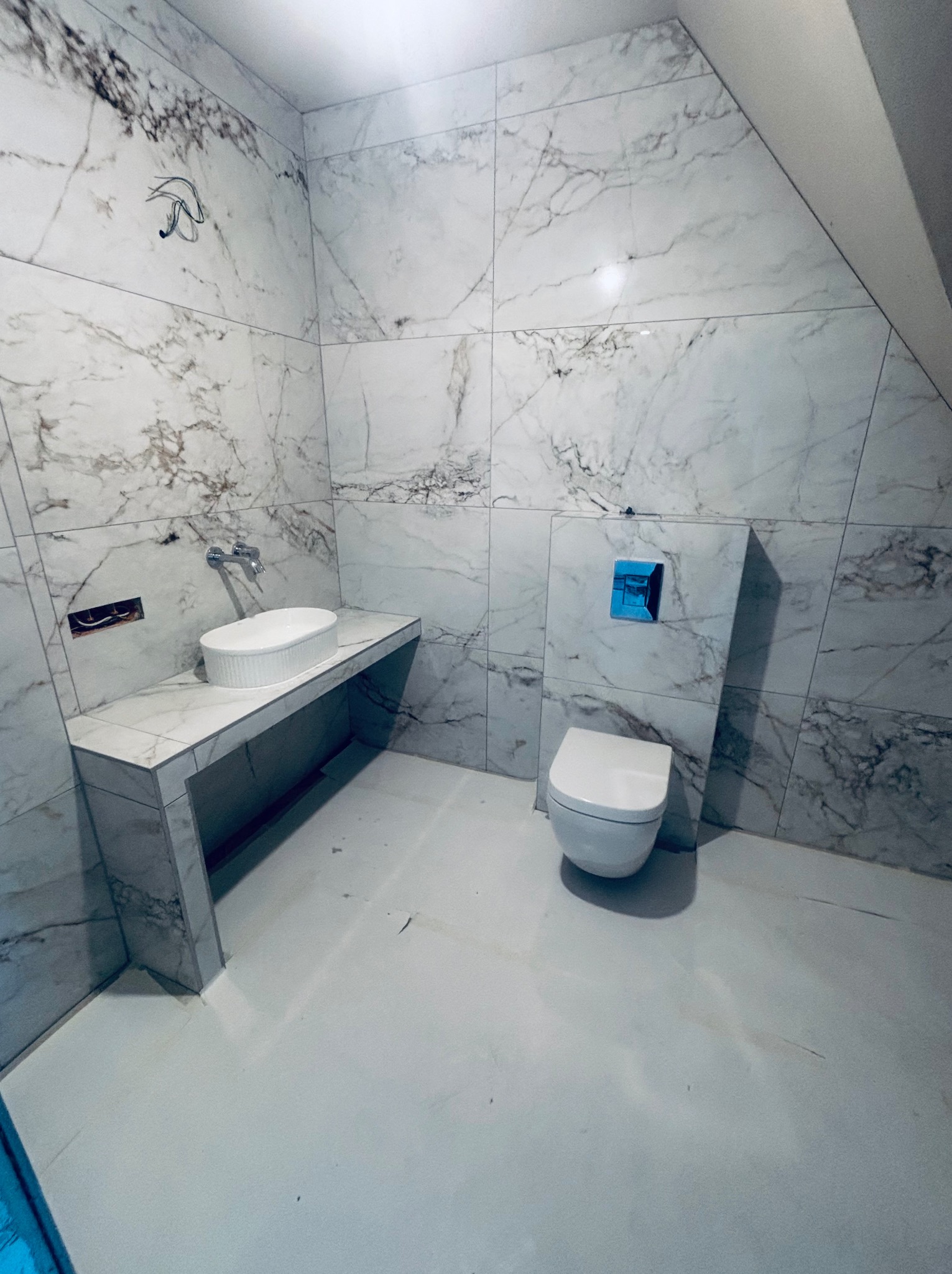 Wykończona łazienka w bieli i marmurze: umywalka nablatowa, toaleta podwieszana z niebieskim przyciskiem, płytki imitujące marmur na ścianach i podłodze.