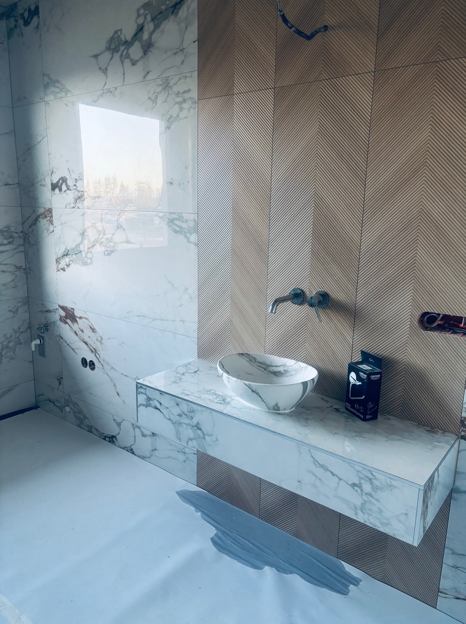 Marmurowa umywalka nablatowa z baterią na tle ściany z płytek w jodełkę. Widoczny fragment podłogi zabezpieczony folią. Nowoczesna aranżacja łazienki w trakcie wykańczania.