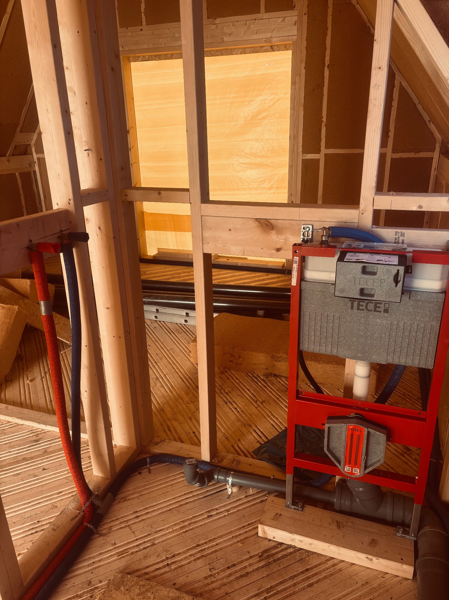 Stelaż podtynkowy TECE zamontowany w drewnianej konstrukcji szkieletowej, z widocznymi rurami doprowadzającymi wodę i odpływem kanalizacyjnym. Surowy stan wykończenia.