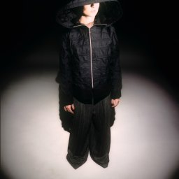 Filip Naumienko Fotografia - Osoba w czarnej kurtce z kapturem, spodnie w paski, oświetlona punktowo na ciemnym tle. Ujęcie od góry podkreśla stylizację.