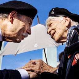Filip Naumienko Fotografia - Zbliżenie na dłonie i twarze starszych osób w mundurach, w beretach, z odznaczeniami, podczas uroczystości, z parasolem w tle i błękitnym niebem.