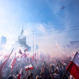 Filip Naumienko Fotografia - Tłum z flagami Polski na tle dymu i Pałacu Kultury, z samolotem na niebie. Ujęcie z dołu, podkreślające skalę wydarzenia i patriotyczny charakter.