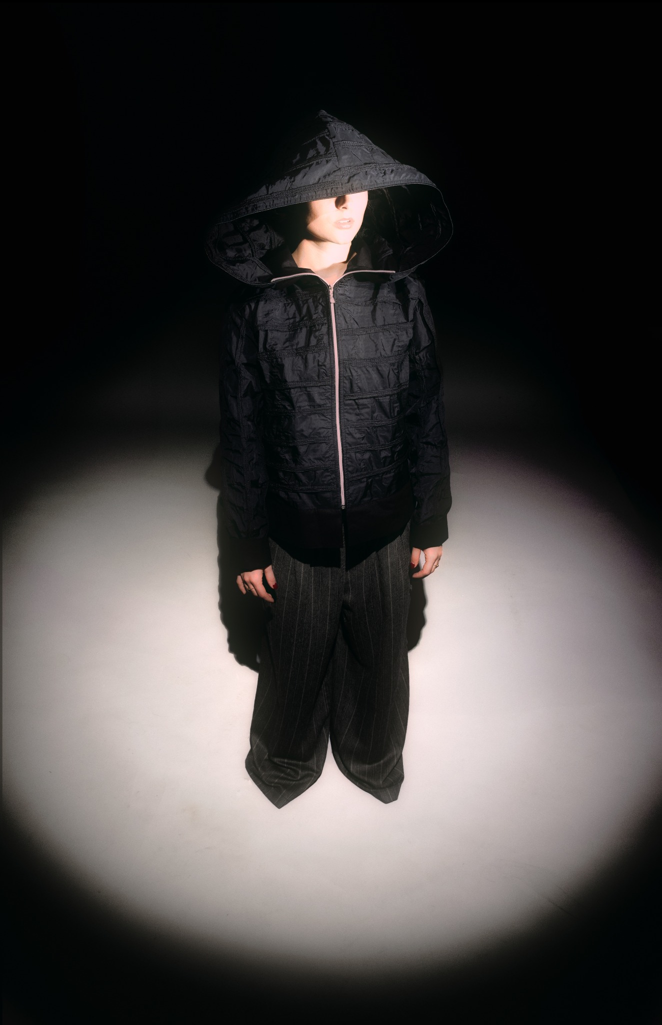 Osoba w czarnej kurtce z kapturem, spodnie w paski, oświetlona punktowo na ciemnym tle. Ujęcie od góry podkreśla stylizację.