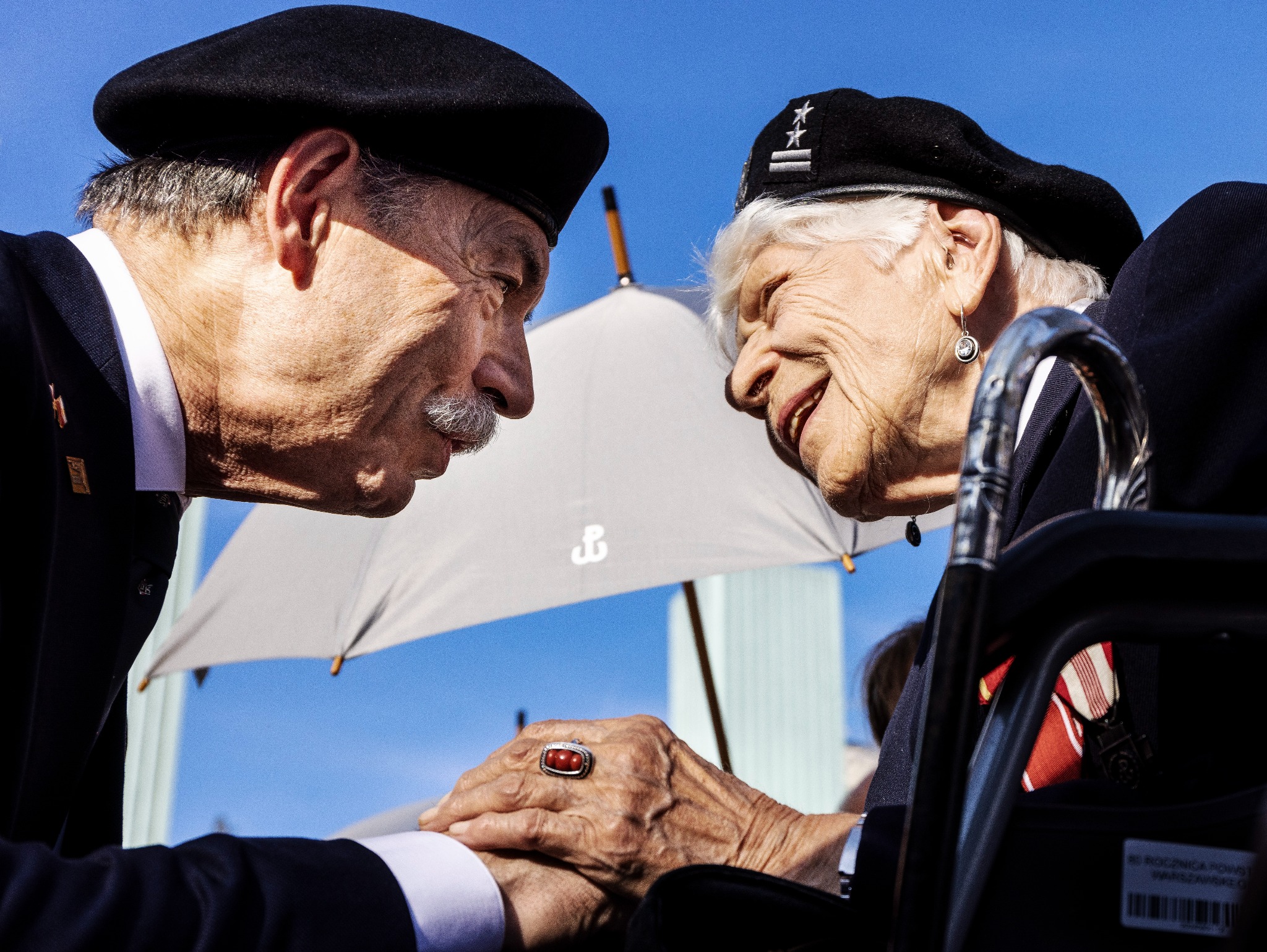 Zbliżenie na dłonie i twarze starszych osób w mundurach, w beretach, z odznaczeniami, podczas uroczystości, z parasolem w tle i błękitnym niebem.