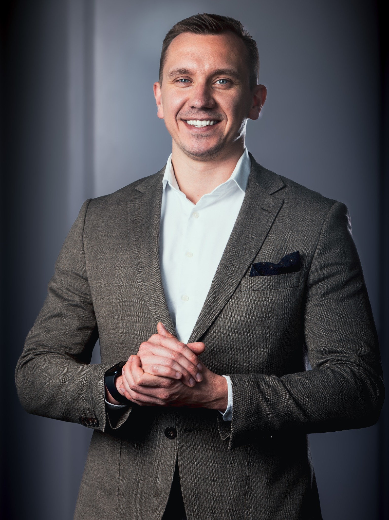 Uśmiechnięty mężczyzna w brązowej marynarce i białej koszuli, dłonie splecione. Portret biznesowy z ciemnym tłem i subtelnym oświetleniem.