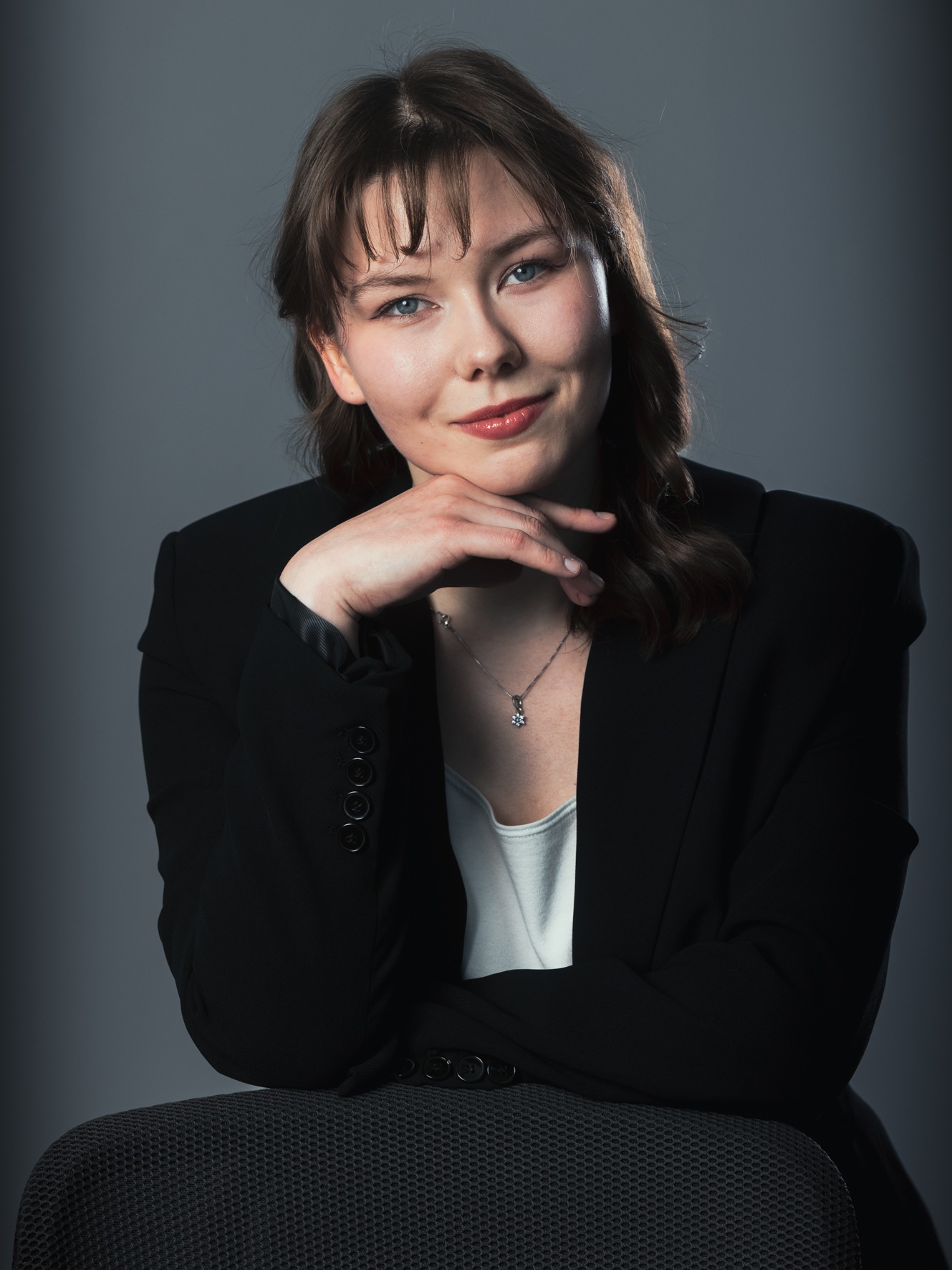 Portret kobiety w czarnej marynarce, opierającej brodę na dłoni, z delikatnym uśmiechem i srebrnym naszyjnikiem na neutralnym, szarym tle. Profesjonalny kadr.