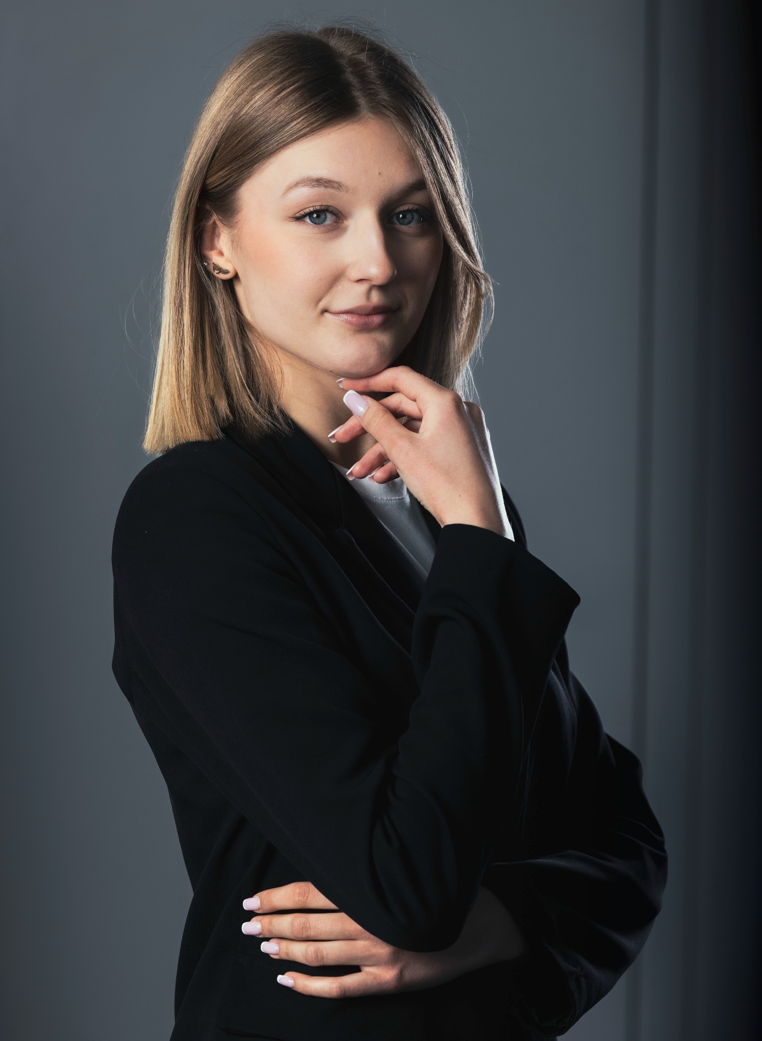 Portret kobiety w czarnej marynarce, z dłonią pod brodą, na szarym tle. Delikatny uśmiech, jasne włosy i subtelny makijaż. Widoczne kolczyki i manicure.