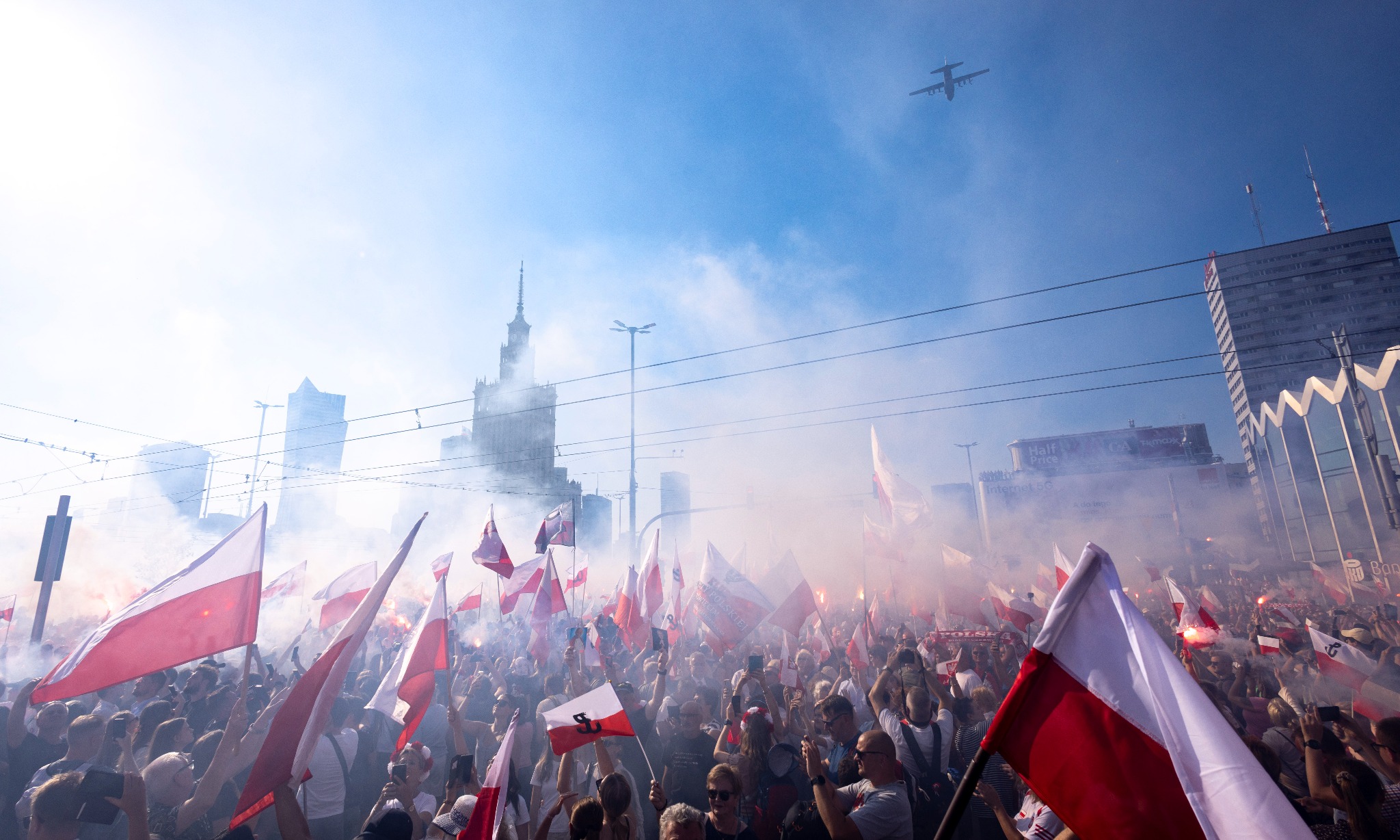 Tłum z flagami Polski na tle dymu i Pałacu Kultury, z samolotem na niebie. Ujęcie z dołu, podkreślające skalę wydarzenia i patriotyczny charakter.