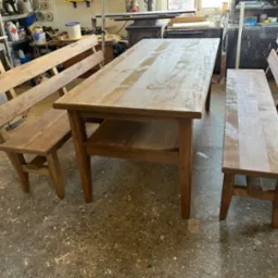 Drewniany stół z dwiema ławkami w warsztacie stolarskim. Naturalne oświetlenie, widoczne ślady obróbki drewna na podłodze, surowe wykończenie mebli.