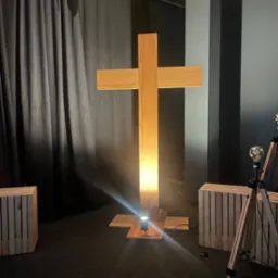 Podświetlony, drewniany krzyż w pomieszczeniu z dekoracjami i czarną zasłoną w tle. Usługi stolarskie w Ustroniu. Nietypowa forma i oświetlenie.