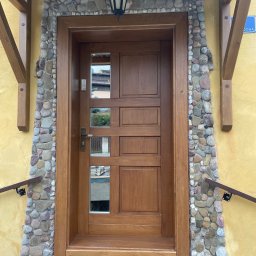 Stolarstwo Adam Pilch - Drewniane drzwi zewnętrzne z szybkami, obramowane kamieniami, pod zadaszeniem. Żółta ściana i drewniane elementy konstrukcyjne. Lampa nad wejściem.