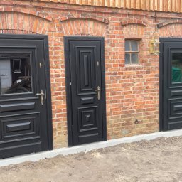 Stolarstwo Adam Pilch - Elewacja z cegły z trzema czarnymi drzwiami. Dwa z przeszkleniami, jeden pełny. Nad środkowymi drzwiami małe okienko i stylizowana lampa.