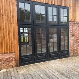 Stolarstwo Adam Pilch - Czarne, przeszklone drzwi zewnętrzne z drewnianą ramą, wkomponowane w elewację z cegły i pionowych desek. Widoczny drewniany taras i drabina w tle.