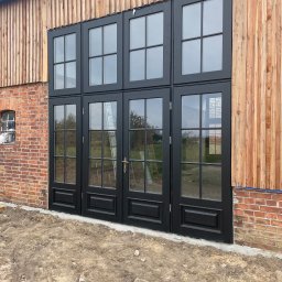 Stolarstwo Adam Pilch - Czarne, przeszklone drzwi zewnętrzne z ramami na tle ceglanej i drewnianej ściany budynku. Widoczna drabina oparta o drzwi. Surowy teren przed budynkiem.