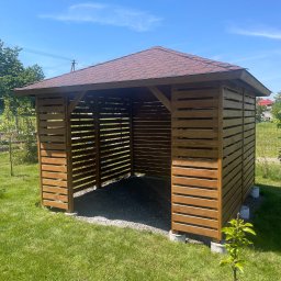 Stolarstwo Adam Pilch - Drewniana altana z brązowym dachem na trawniku w słoneczny dzień. Konstrukcja z poziomych desek, widoczne elementy betonowe podstawy. Błękitne niebo w tle.