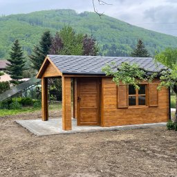 Stolarstwo Adam Pilch - Brązowa, drewniana altana z szarym, gontowym dachem na tle zielonego wzgórza. Otwarta przestrzeń z kolumną wejściową i otwarte drzwi.