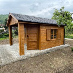 Stolarstwo Adam Pilch - Drewniana altana z otwartym gankiem i oknem, pomalowana na brązowo, stoi na wyłożonym kostką placu. W tle zielone wzgórza i drzewa.