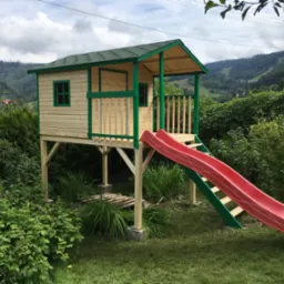 Drewniany domek dla dzieci na podwyższeniu z czerwoną zjeżdżalnią, otoczony zielenią. Widok na góry w tle, dom z zielonymi akcentami.