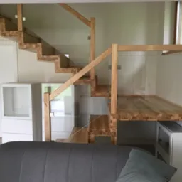 Nowoczesne schody drewniane z szklaną balustradą, widok z dołu. Stopnie z litego drewna, minimalistyczny design, jasne wnętrze. Widoczna część sofy.