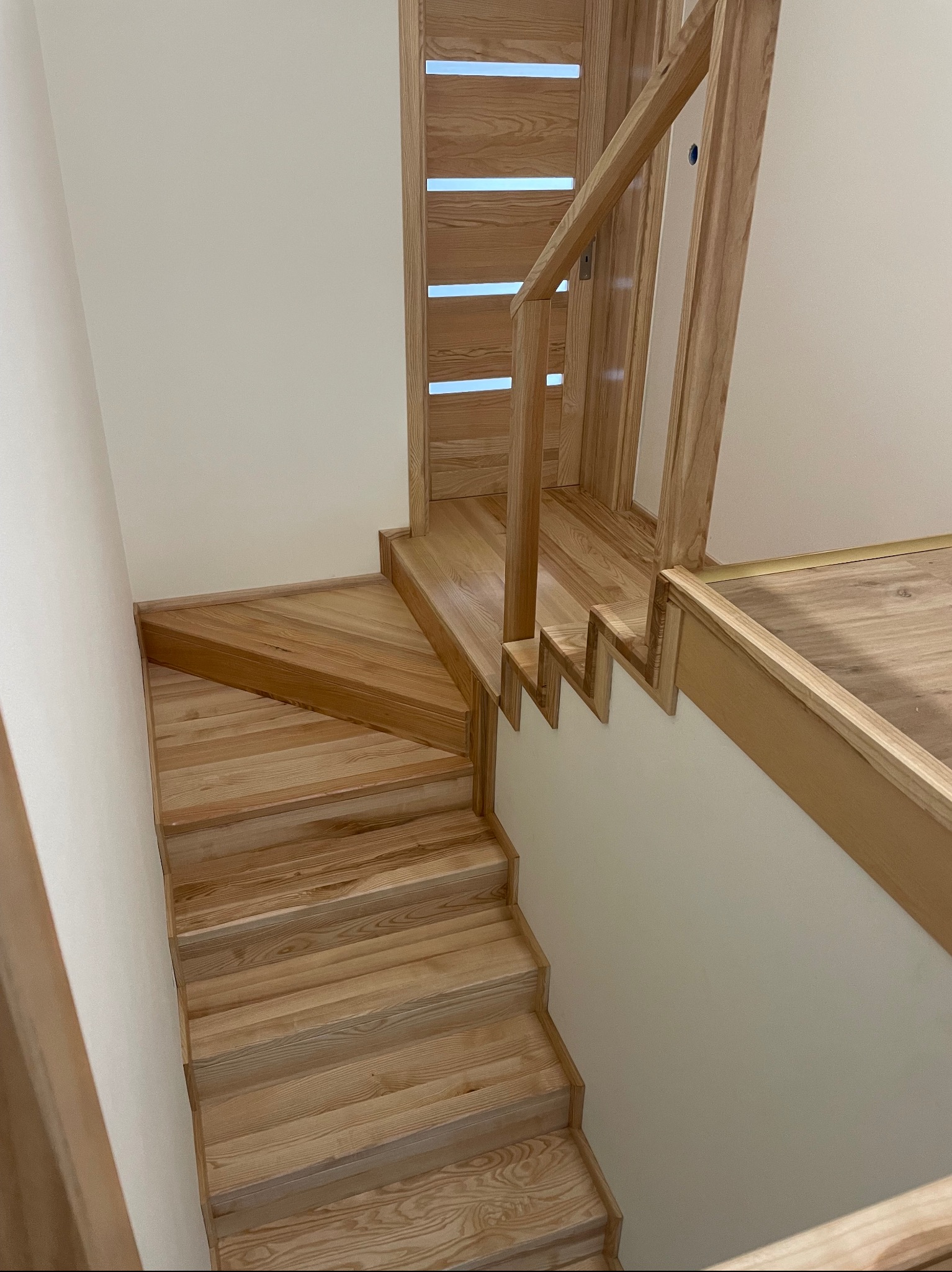 Jasne, drewniane schody z balustradą, z widocznym fragmentem drzwi z poziomymi przeszkleniami, prowadzące do górnej kondygnacji. Widok z góry.