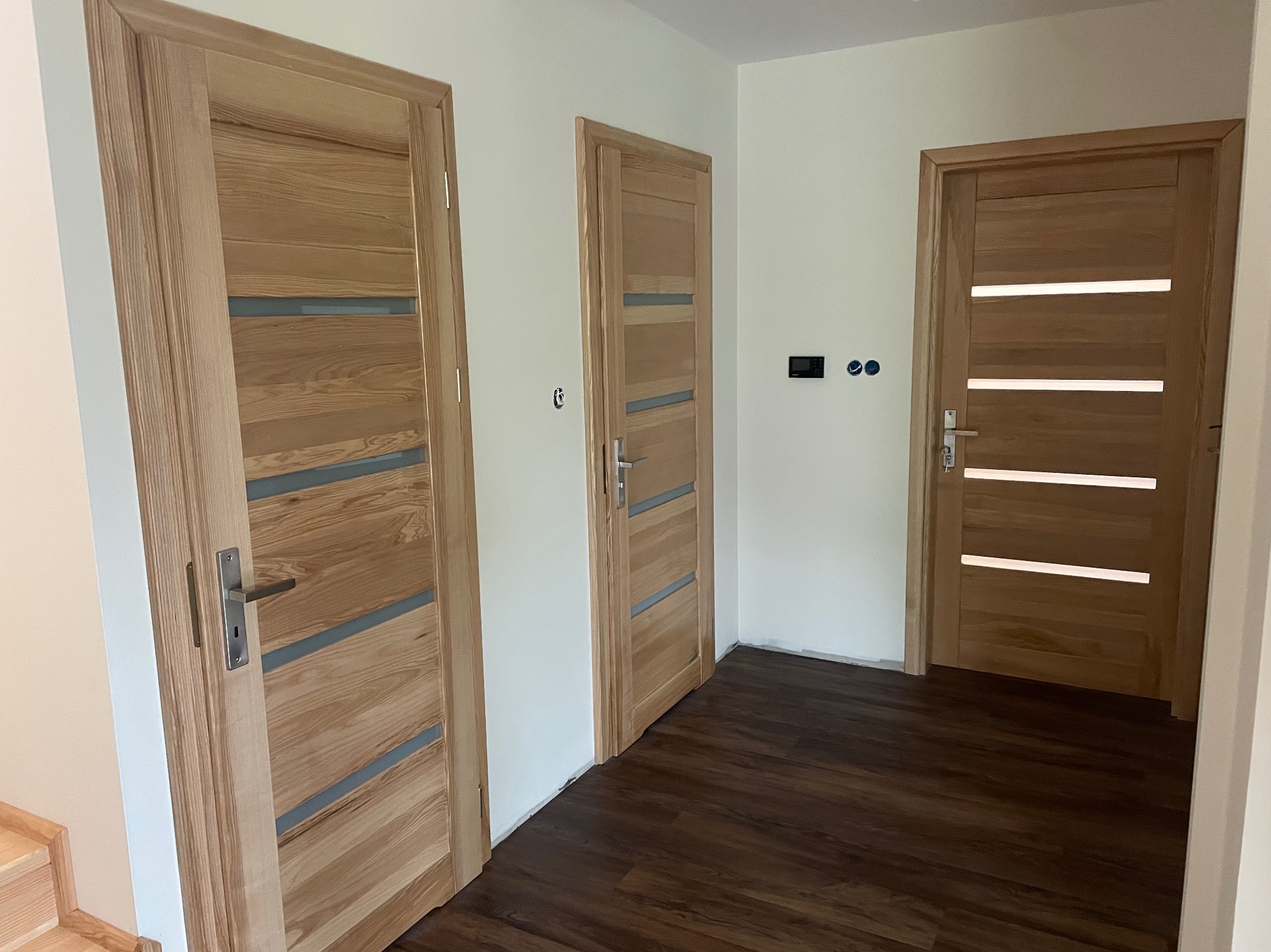 Jasne, drewniane drzwi wewnętrzne z przeszkleniami, widoczne z perspektywy wnętrza. Brązowa podłoga, białe ściany, trzy skrzydła drzwiowe w różnych odległościach.