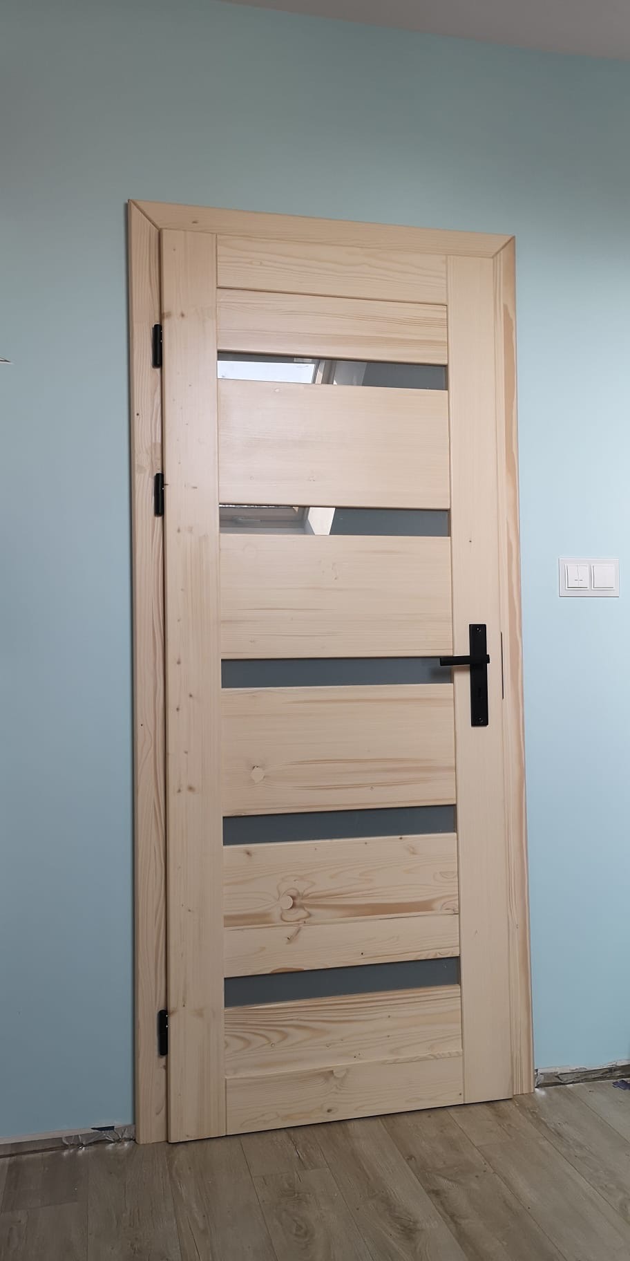 Surowe, drewniane drzwi wewnętrzne z czterema poziomymi przeszkleniami, czarna klamka, zawieszone na tle błękitnej ściany. Widoczne detale wykończenia i surowość drewna.