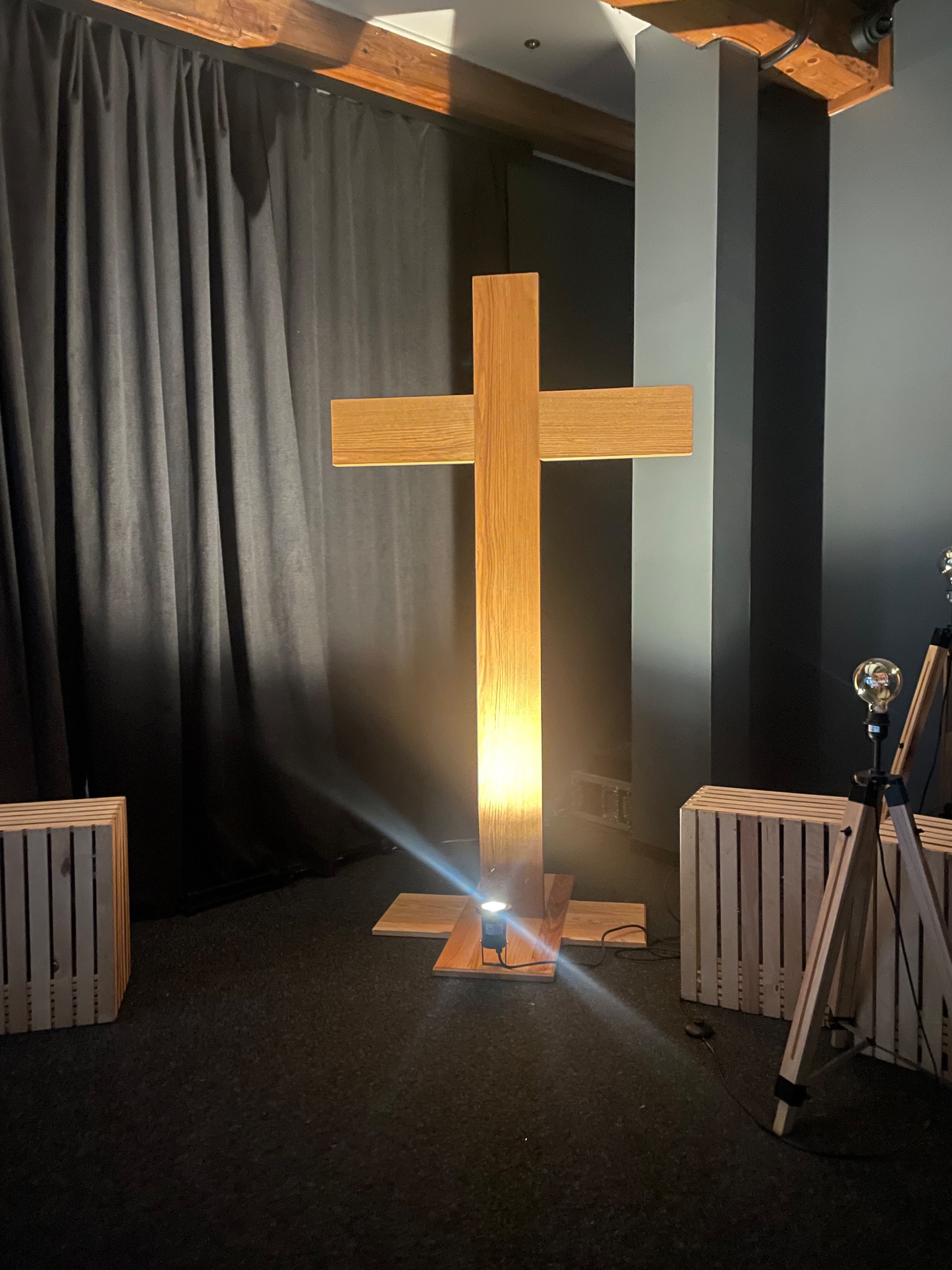 Podświetlony, drewniany krzyż w pomieszczeniu z dekoracjami i czarną zasłoną w tle. Usługi stolarskie w Ustroniu. Nietypowa forma i oświetlenie.