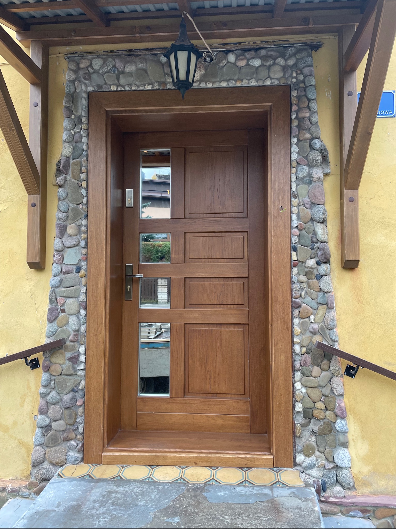 Drewniane drzwi zewnętrzne z szybkami, obramowane kamieniami, pod zadaszeniem. Żółta ściana i drewniane elementy konstrukcyjne. Lampa nad wejściem.
