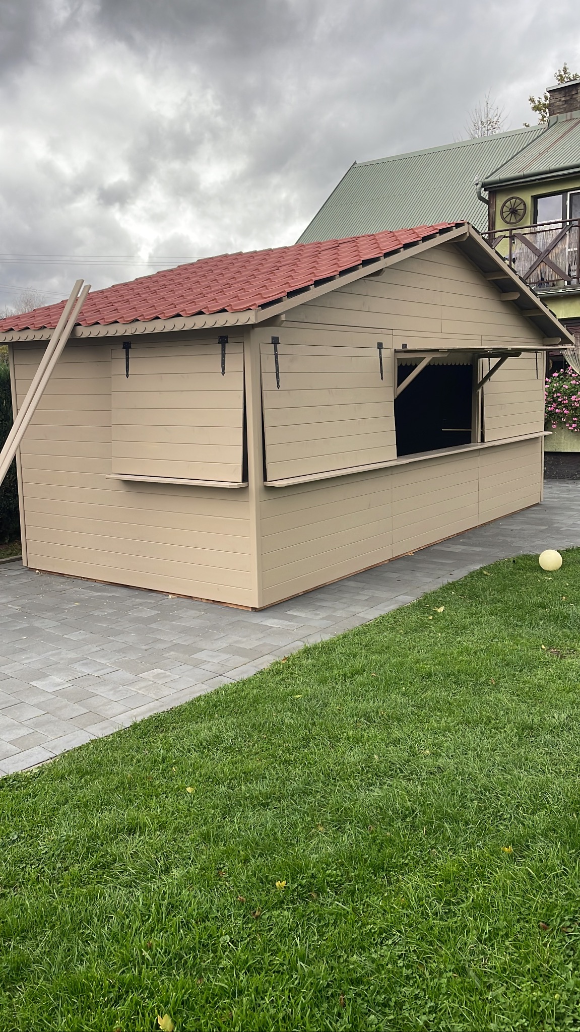 Beżowa altana z czerwoną dachówką na trawniku, z otwartymi okienkami. Drabina oparta o bok. Na drugim planie dom z balkonem i dekoracyjnym kołem.