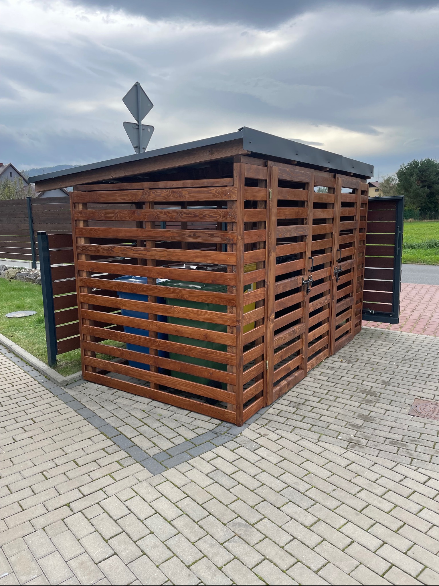 Drewniana zabudowa śmietnikowa z dwoma pojemnikami w środku, wykonana z brązowych desek, z szarym dachem, zamykana na zatrzask, na tle kostki brukowej i pochmurnego nieba.
