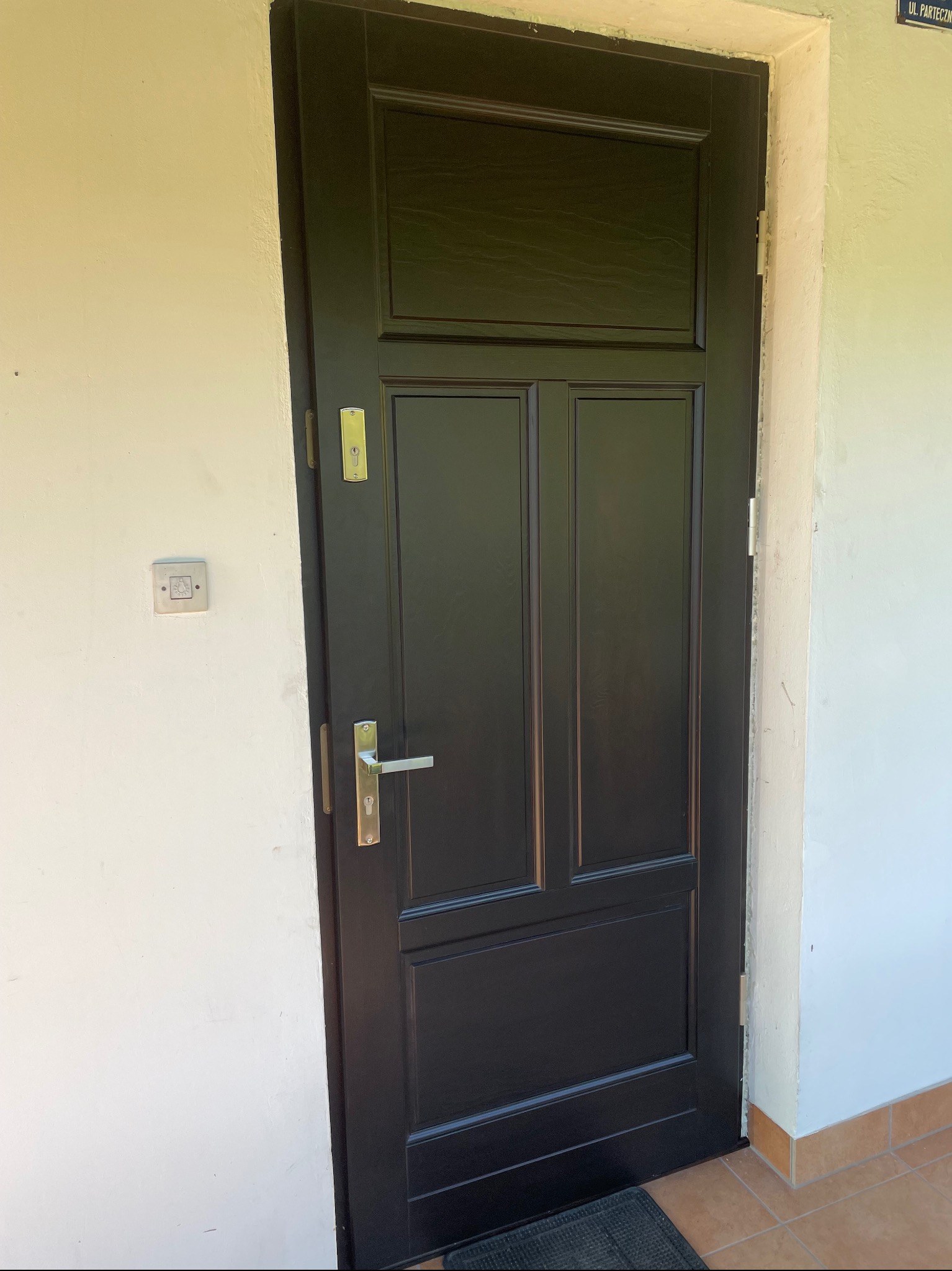 Ciemne drzwi zewnętrzne z panelem i srebrną klamką na tle białej ściany. Wykończenie z płytkami. Widoczna tabliczka z nazwą ulicy.