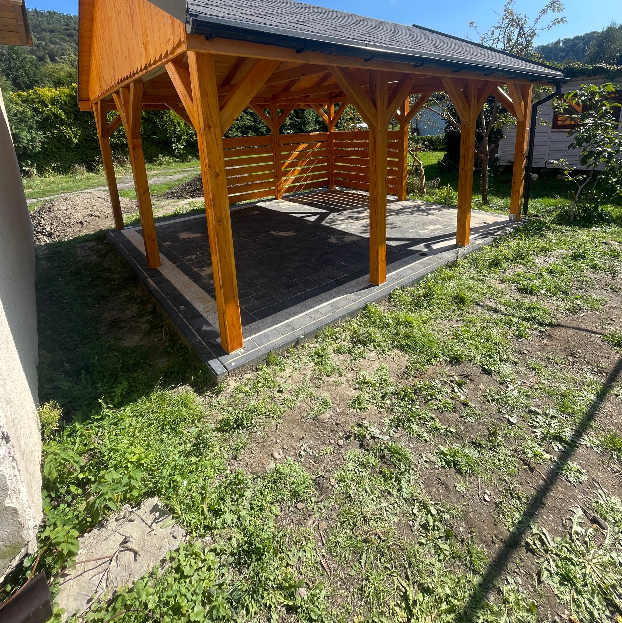 Drewniana altana z czarną dachówką i brukowaną podłogą, otoczona zielenią. Widok z zewnątrz, w słoneczny dzień. Klasyczna konstrukcja z poziomymi deskami w tylnej części.