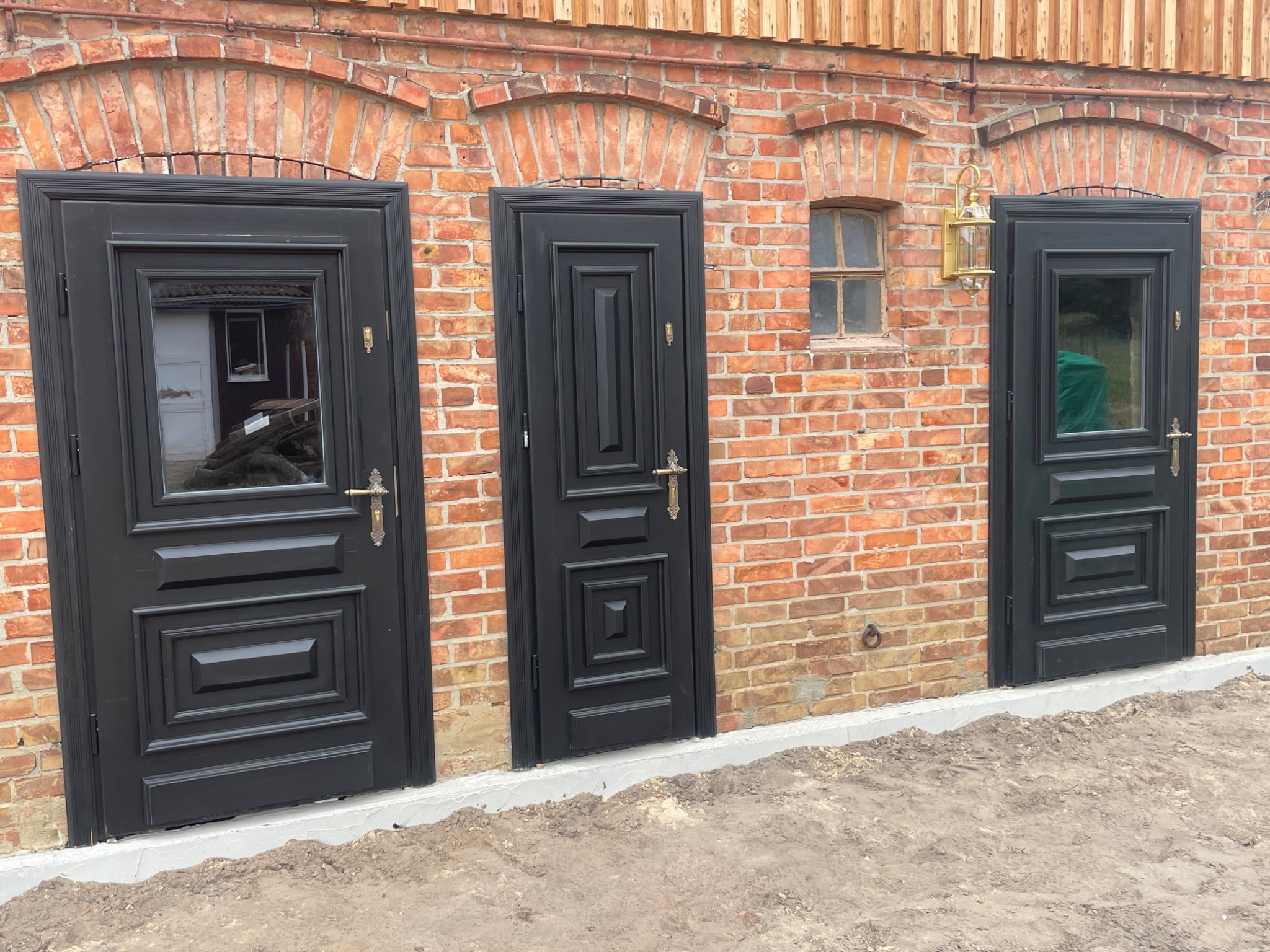 Elewacja z cegły z trzema czarnymi drzwiami. Dwa z przeszkleniami, jeden pełny. Nad środkowymi drzwiami małe okienko i stylizowana lampa.