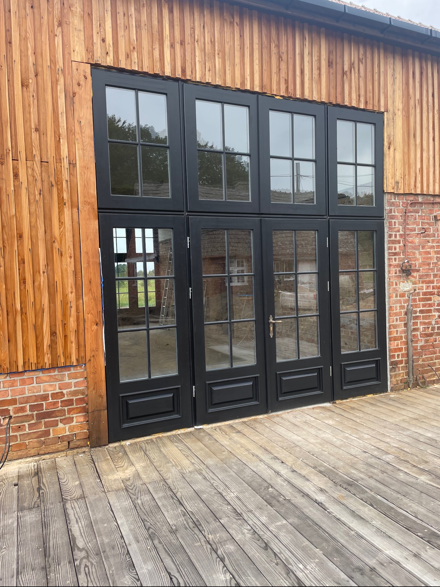 Czarne, przeszklone drzwi zewnętrzne z drewnianą ramą, wkomponowane w elewację z cegły i pionowych desek. Widoczny drewniany taras i drabina w tle.