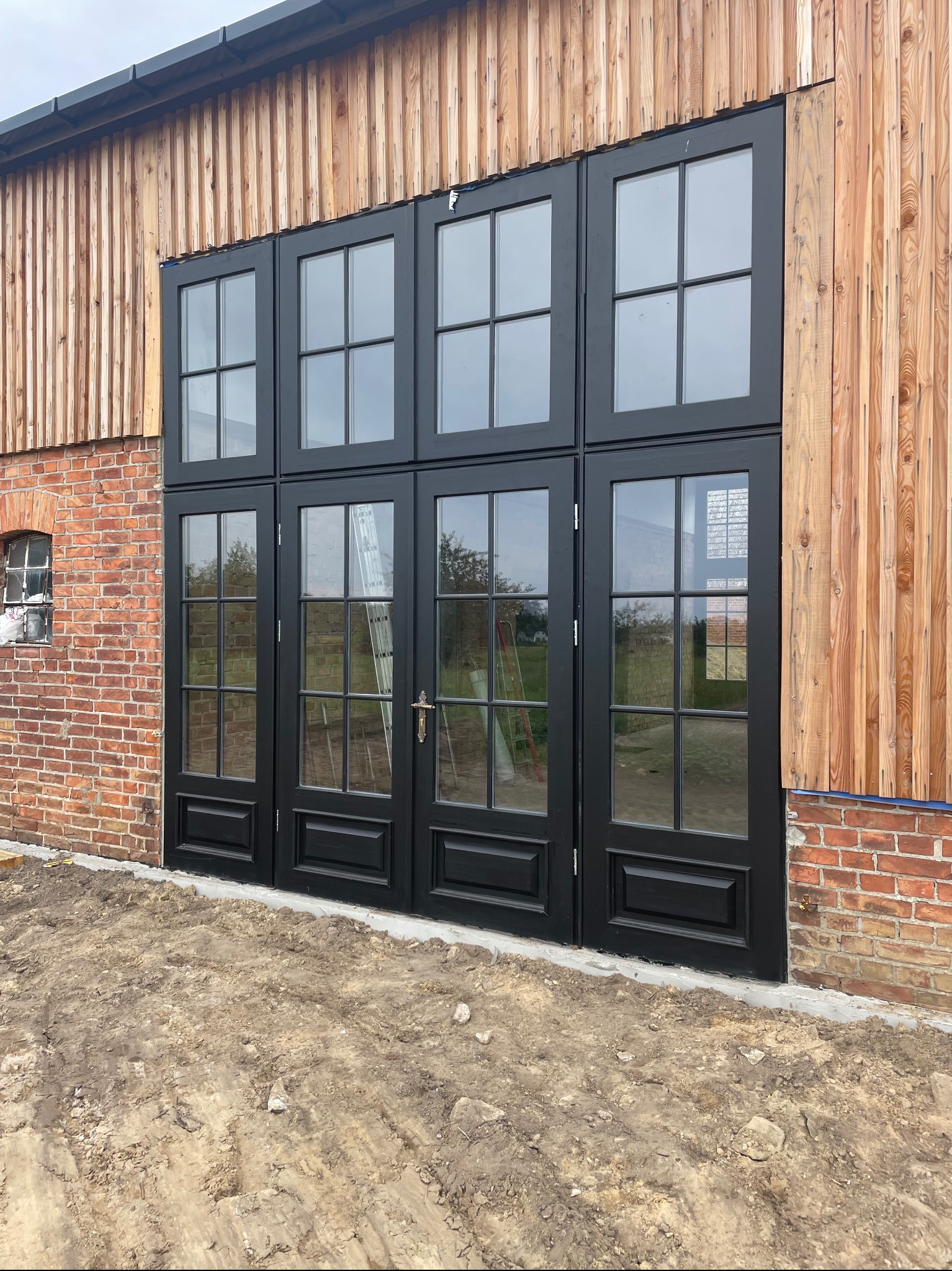 Czarne, przeszklone drzwi zewnętrzne z ramami na tle ceglanej i drewnianej ściany budynku. Widoczna drabina oparta o drzwi. Surowy teren przed budynkiem.