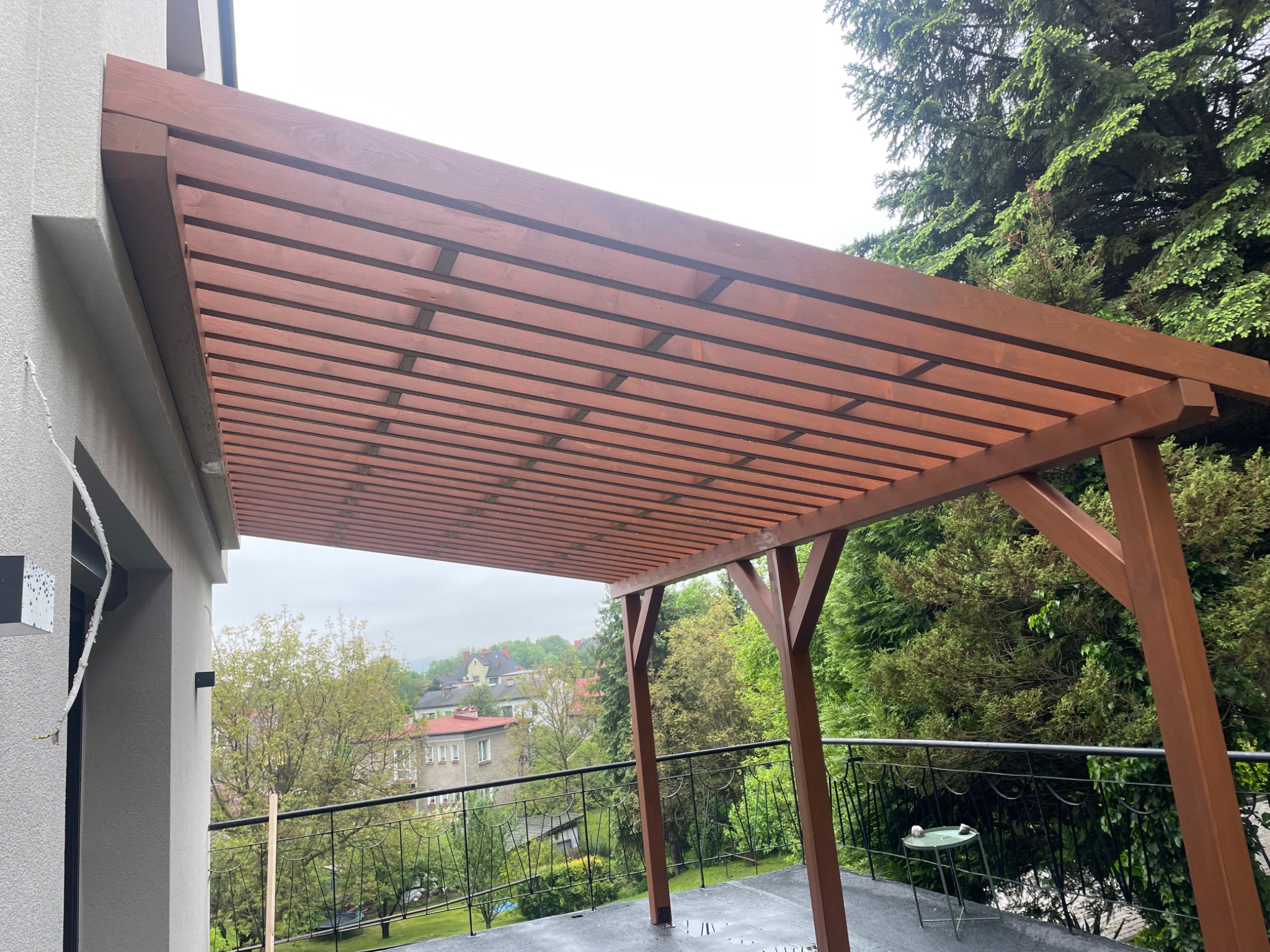 Brązowa pergola z drewna na balkonie z widokiem na zieleń i zabudowę miejską w tle. Konstrukcja przylega do ściany budynku z lewej strony.